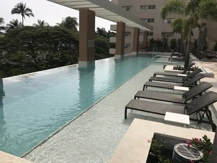 Infinity-edge pool with a shallow tiled sun shelf and dark loungers set along the edge beneath a concrete colonnade